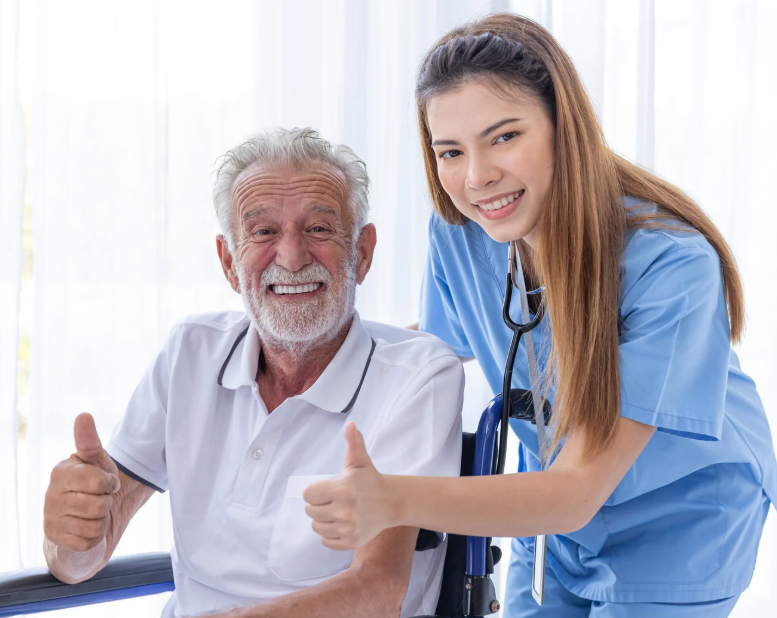 Caregiver in blue scrubs with an older adult in a wheelchair, both smiling and giving thumbs up